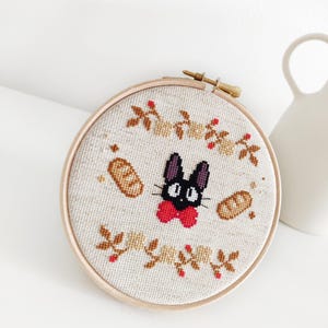 May include: A black cat with a red bow tie and a white face is embroidered on a beige fabric in a round wooden hoop. The cat is surrounded by brown bread and small brown leaves with red berries.
