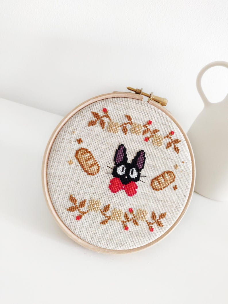 May include: A black cat with a red bow tie and a white face is embroidered on a beige fabric in a round wooden hoop. The cat is surrounded by brown bread and small brown leaves with red berries.