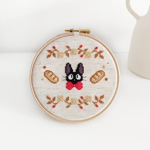 May include: A black cat with a red bow tie embroidered on a beige fabric with a brown wooden hoop. The cat is surrounded by brown bread and red berries.