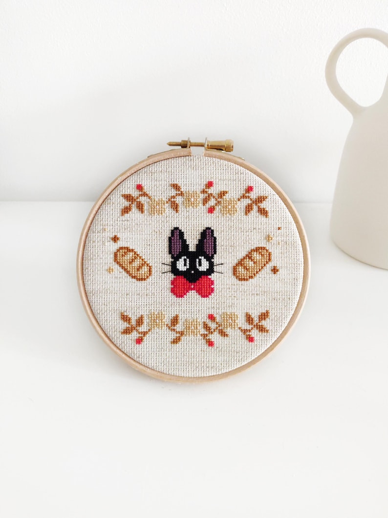 May include: A black cat with a red bow tie embroidered on a beige fabric with a brown wooden hoop. The cat is surrounded by brown bread and red berries.