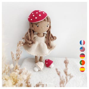 Peut inclure: Une poupée en crochet portant un chapeau de champignon rouge et blanc et une robe crème. La poupée tient un petit champignon blanc dans une main et un champignon rouge dans l'autre. La poupée est assise sur une surface en tissu blanc.