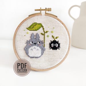 May include: A cross-stitch embroidery in a wooden hoop features a gray Totoro character with a green leaf and a black soot sprite. The design is on a beige fabric. The text "PDF PATTERN" is in a circle at the bottom.