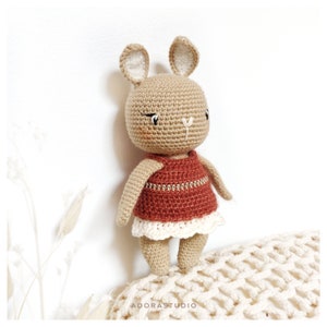 Peut inclure: Un jouet de lapin en crochet portant une robe rouge et beige avec une bordure en dentelle blanche. Le lapin a des yeux bruns et un corps beige.