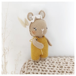 Peut inclure: Un animal en peluche crocheté en forme de lapin portant un bandeau crocheté blanc et une salopette crocheté jaune. Le lapin tient une petite brindille dans sa main.