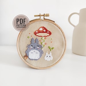 May include: A cross-stitch embroidery hoop with a gray Totoro character, a white bunny, and a red mushroom with white spots. The embroidery is on a beige fabric.