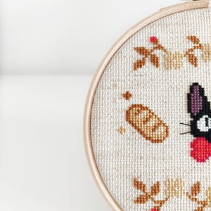 May include: A black cat with a red bow, a loaf of bread, and floral embroidery on a white background. The embroidery is in shades of brown, beige, and red. The cat is a popular character from the animated film "My Neighbor Totoro".