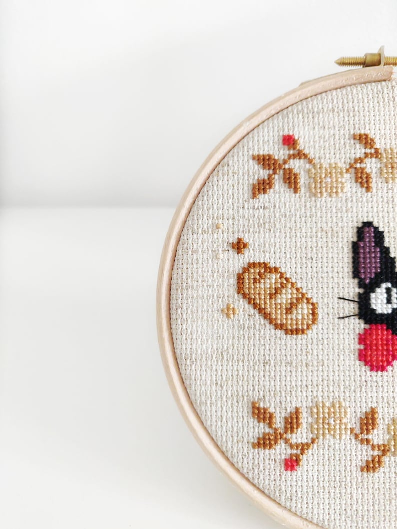 May include: A black cat with a red bow, a loaf of bread, and floral embroidery on a white background. The embroidery is in shades of brown, beige, and red. The cat is a popular character from the animated film "My Neighbor Totoro".