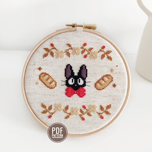 Puede incluir: Un patrón de punto de cruz con un gato negro con una pajarita roja, rodeado de pan y detalles florales. El patrón está enmarcado en un aro de madera y tiene la etiqueta "PDF PATTERN".