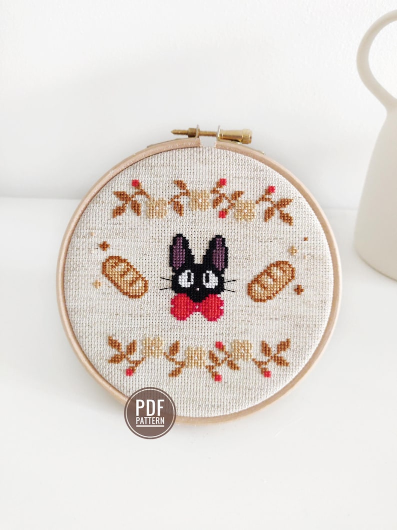 May include: A cross-stitch pattern featuring a black cat with a red bow tie, surrounded by bread and floral accents. The pattern is framed in a wooden hoop and labeled "PDF PATTERN".