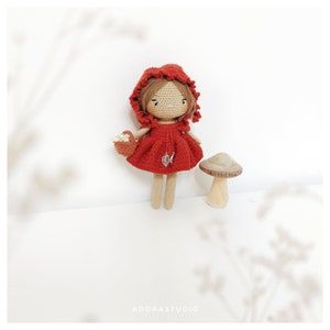 May include: A crocheted doll wearing a red hooded dress and holding a basket of flowers. The doll is standing next to a small wooden mushroom.