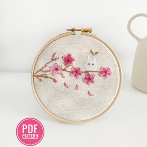 Peut inclure: Un motif de point de croix représentant un lapin blanc avec des fleurs de cerisier roses sur une branche. Le motif est encadré dans un cercle en bois.