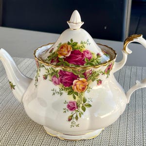 May include: A white porcelain teapot with a lid, decorated with a floral rose pattern in pink, yellow, and red. The teapot has gold detailing on the handle, spout, and lid. A classic tea set item.