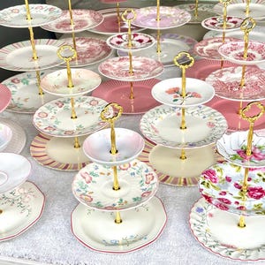 Può includere: Una collezione di alzatine a più livelli con manici dorati. Le alzatine presentano piatti con vari motivi floreali e a righe, con schemi di colori rosa e bianco. Queste alzatine decorative sono ideali per dolci o antipasti.