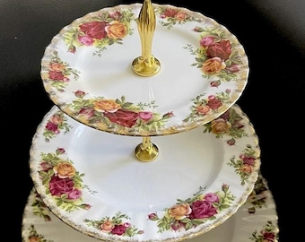 Peut inclure: Présentoir à gâteaux à trois niveaux avec une poignée et des supports dorés. Chaque assiette blanche est ornée d'un motif floral de roses rouges, roses et jaunes. Les bords des assiettes sont festonnés et dorés.