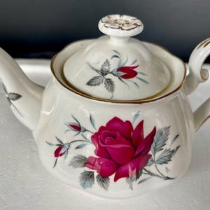 May include: Cream-coloured porcelain teapot featuring a large red rose and smaller buds. The teapot has a gold trim and a matching lid with a floral design. A classic tea set item.