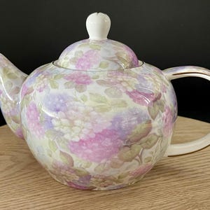 Op de afbeelding: Een witte porseleinen theepot met een bloemenpatroon in tinten roze, paars en groen. De theepot heeft een deksel en een handvat.
