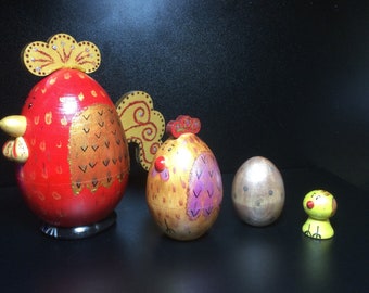 Könnte beinhalten: Ein Set aus vier dekorativen Holzfiguren, bestehend aus einem großen roten Hahn mit goldenen Akzenten, einer mittelgroßen goldenen Henne mit rosa Details, einer metallisch silbernen eiförmigen Figur und einem kleinen gelben Küken mit rotem Schnabel.