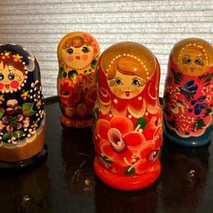 May include: Four Russian nesting dolls, each with a unique painted design. The dolls feature floral patterns, and the faces are painted with rosy cheeks and detailed features. The color palette includes blues, reds, yellows, and greens.