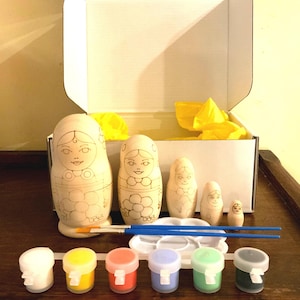 May include: A DIY painting kit featuring unfinished wooden nesting dolls, paint pots in various colors, paintbrushes, and a palette. The dolls are in a box with yellow tissue paper. The kit is designed for creative crafting.