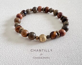 May include: A beaded bracelet featuring a mix of wooden, tiger eye, and metal beads in shades of brown, gold, and bronze. The bracelet has a stretch design and is displayed on a textured white surface. The brand name "CHANTILLY" is visible.