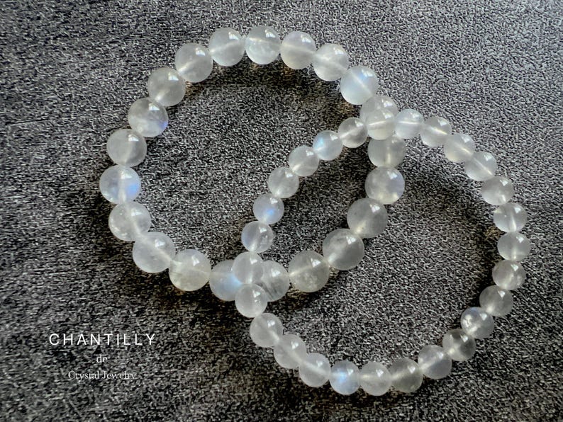 May include: Two white moonstone bracelets with a shimmery iridescent finish. The bracelets are made of round beads and are stacked on top of each other.