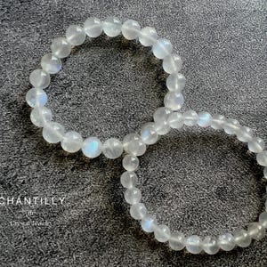 May include: Two white moonstone beaded bracelets on a gray textured surface. The bracelets are made of round beads and are stacked on top of each other.