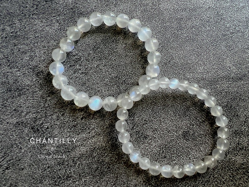 May include: Two white moonstone beaded bracelets on a gray textured surface. The bracelets are made of round beads and are stacked on top of each other.