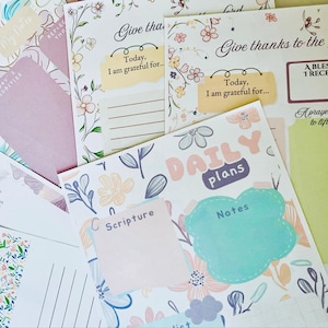 May include: A selection of floral-patterned stationery, including notepads and cards. The designs feature pastel colours, botanical illustrations, and handwritten text such as "Give thanks to the Lord" and "Daily Plans".