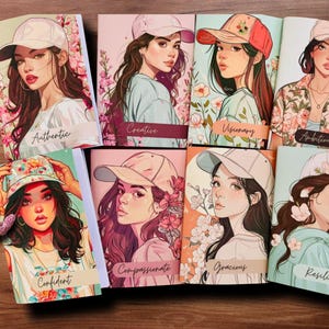 Op de afbeelding: Een verzameling notitieboekjes met illustraties van vrouwen met baseballpetten. Elk notitieboekje heeft een ander woord, waaronder "Authentiek", "Creatief", "Visionair", "Zelfverzekerd", "Mededogend", "Gracieus", "Veerkrachtig" en "Ambitieus".