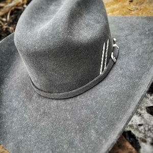 Op de afbeelding: Een grijze wollen cowboyhoed met een zilveren band en decoratieve accenten. De hoed ligt op een rustiek houten oppervlak.