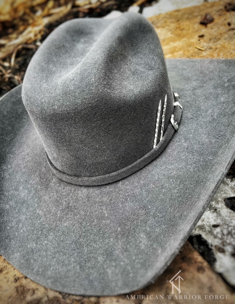 Op de afbeelding: Een grijze wollen cowboyhoed met een zilveren band en decoratieve accenten. De hoed ligt op een rustiek houten oppervlak.