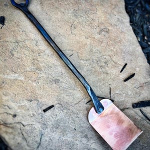 Peut inclure: Une spatule en métal forgée à la main avec une lame en cuivre et une poignée en anneau. La spatule repose sur une surface en pierre texturée.