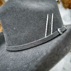 Op de afbeelding: Een grijze wollen cowboyhoed met een leren band en twee zilveren hoedspelden. De hoed ligt op een wit oppervlak.