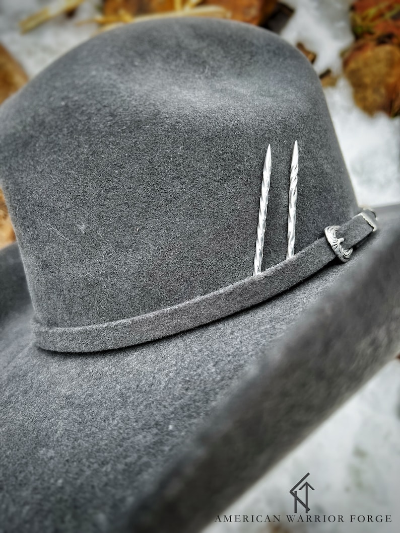 Op de afbeelding: Een grijze wollen cowboyhoed met een leren band en twee zilveren hoedspelden. De hoed ligt op een wit oppervlak.