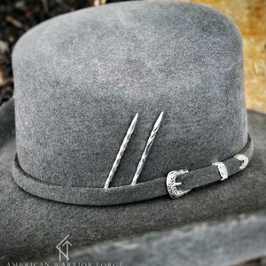 Op de afbeelding: Een grijze wollen cowboyhoed met een zilveren gesp en twee zilveren pinnen. De hoed ligt op een bruine ondergrond. American Warrior Forge is onderaan de afbeelding gedrukt.