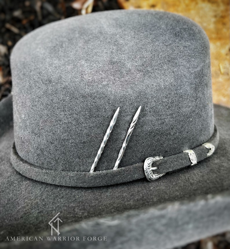 Op de afbeelding: Een grijze wollen cowboyhoed met een zilveren gesp en twee zilveren pinnen. De hoed ligt op een bruine ondergrond. American Warrior Forge is onderaan de afbeelding gedrukt.