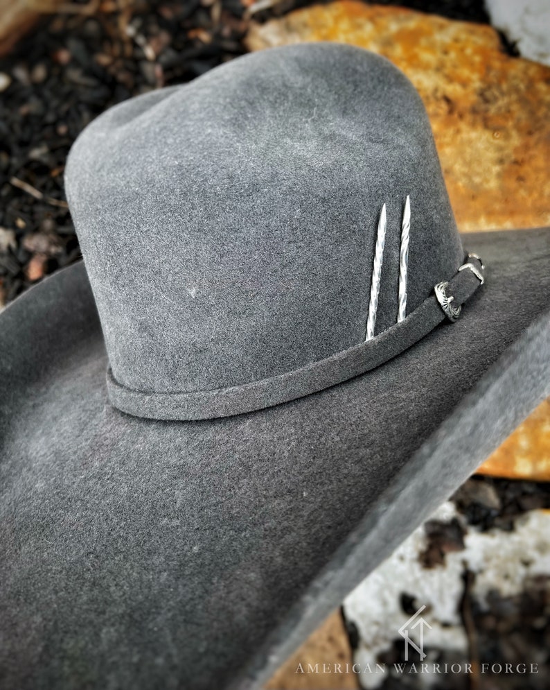 Op de afbeelding: Een grijze wollen cowboyhoed met een leren band en zilveren accenten. De hoed heeft een brede rand en een hoge kroon.