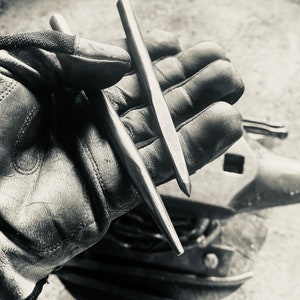 Peut inclure: Deux poinçons métalliques sont tenus dans une main gantée, avec une enclume en arrière-plan. Les poinçons sont longs, fins et ont une extrémité pointue. L'image est en noir et blanc, mettant en évidence les outils utilisés en métallurgie.