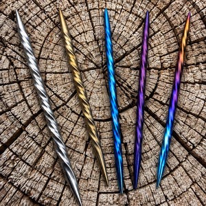 May include: Five metal hair sticks with a twisted spiral design. The sticks are finished in metallic silver, gold, blue, purple, and a multi-colored gradient of blue, purple, and orange. Each stick features a pointed tip and a tapered end.