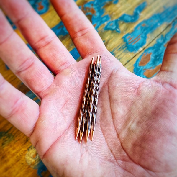 May include: Four twisted metal knitting needles, with a copper finish, are held in a hand. The needles are about 4 inches long.