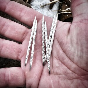 Op de afbeelding: Een set van zes zilveren, gedraaide, puntige metalen staven, vastgehouden in een hand. De staven zijn ongeveer 10 cm lang.