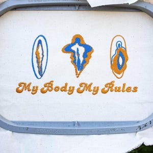 May include: An embroidery hoop holds a cream-colored fabric with three abstract blue and gold embroidered shapes. The text "My Body My Rules" is embroidered in gold thread below the shapes. The hoop is gray.