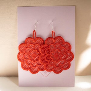 May include: A pair of red crocheted earrings with a floral design. The earrings are displayed on a pink card.
