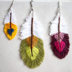 Puede incluir: Tres pendientes de plumas hechos a mano. Cada pendiente presenta una parte superior blanca con un tallo central marrón y una parte inferior colorida. Uno es amarillo con un acento azul, otro es verde y naranja, y el último es rosa y burdeos.