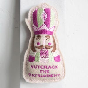 Może przedstawiać: Haftowana naszywka przedstawiająca dziadka do orzechów w różowo-zielonym kapeluszu i mundurze. Pod spodem wyhaftowano napis "Nutcrack the Patriarchy". Naszywka ma kremową obwódkę.