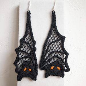 May include: A pair of black bat-shaped earrings. The earrings are made of a delicate, open-weave material, with a bat silhouette. The bat's eyes are embroidered in orange. The earrings hang from silver-colored hooks.