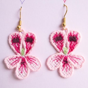 May include: A pair of handmade earrings with a floral design. The earrings are made of light pink openwork lace, with dark pink and brown detailing. A small green accent is in the centre. The earrings have gold-coloured ear wires.