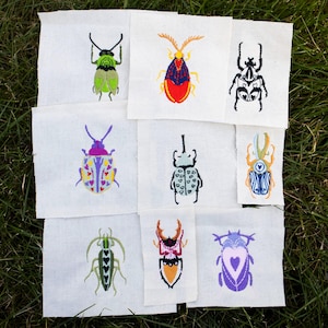 May include: Nine embroidered insect patches on white fabric. The insects are in various colors, including green, red, blue, and purple, with detailed stitching. The patches are arranged on a bed of green grass.