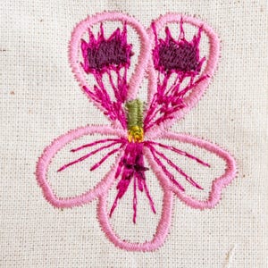 May include: Embroidered floral design in shades of pink and purple on a cream-colored fabric. The flower features a detailed outline with delicate stitching, showcasing a central cluster of dark purple petals and a green stem.