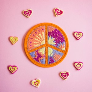 Puede incluir: Un parche con el símbolo de la paz con un diseño bordado de colores de un sol, flores y manos. El parche está rodeado de diez pequeños parches en forma de corazón de color amarillo y rosa.